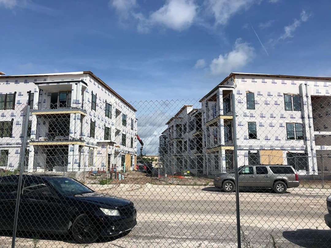 Construction site of two multi-unit buildings with a chain-link fence and vehicles parked nearby.