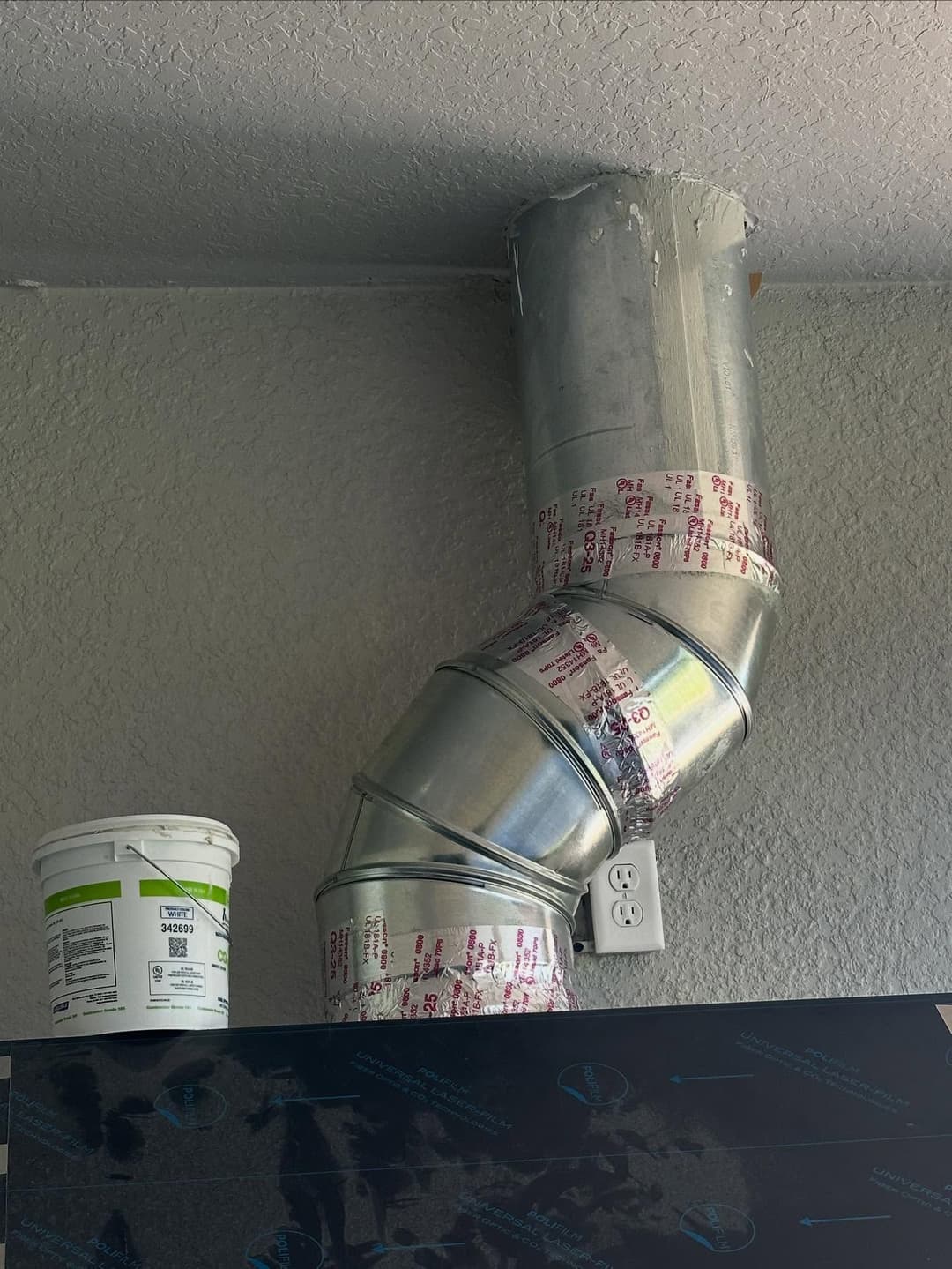 Metal ductwork with a 90-degree bend, taped, above a paint bucket and electrical outlet.