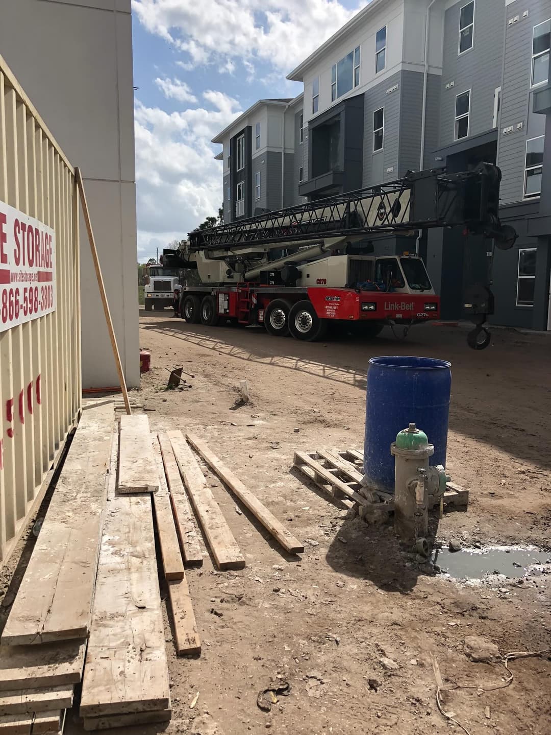 Construction site with a crane, storage container, and wooden planks near apartment buildings.